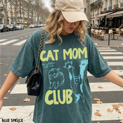A woman wearing an oversized slate blue-green graphic t-shirt with 'CAT MOM CLUB' printed in yellow and light blue, styled with a beige cap and a black quilted shoulder bag on a city street.