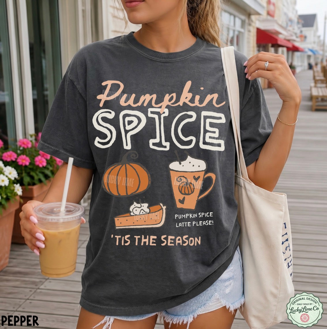 A woman wearing a dark charcoal gray graphic t-shirt with 'Pumpkin Spice' written in peach and white text, featuring illustrations of a pumpkin, latte, and pie. She is holding an iced coffee and carrying a canvas tote bag on a wooden boardwalk.