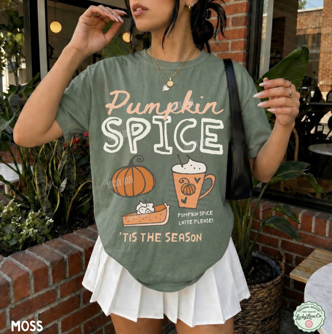A woman wearing a sage green graphic t-shirt with 'Pumpkin Spice' text and autumn illustrations, paired with a white pleated skirt, standing in front of a brick patio storefront.