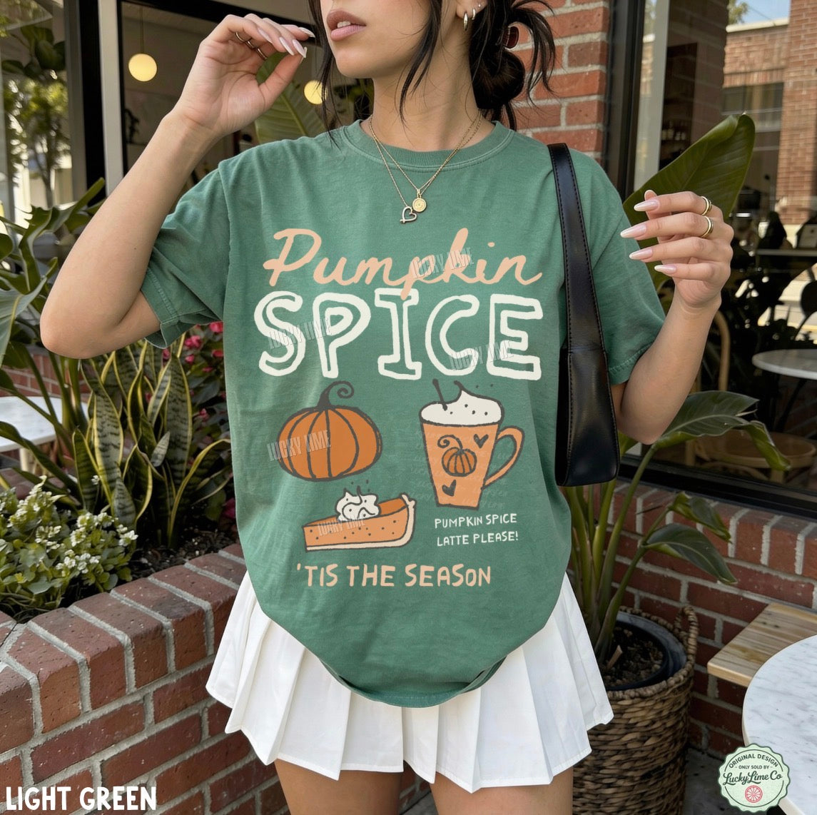 A woman wearing a light green graphic t-shirt with 'Pumpkin Spice' and autumn illustrations, paired with a white pleated skirt, standing outdoors in front of a brick wall and plants.