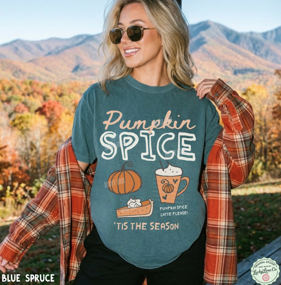 A woman outdoors wearing a teal graphic T-shirt that reads 'Pumpkin Spice' with autumn-themed illustrations, paired with an open red-orange plaid flannel shirt. Mountains and fall foliage are visible in the background.
