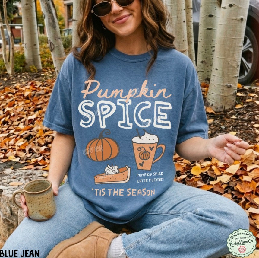 A woman sitting outdoors in autumn trees wears a blue graphic T-shirt that reads 'Pumpkin Spice' with fall-themed illustrations, paired with light blue jeans and brown boots. She holds a mug, and the background shows fallen leaves and birch trees.
