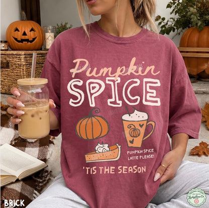 A woman wearing a maroon graphic t-shirt with 'Pumpkin Spice' text and fall-themed illustrations, holding an iced coffee in a cozy autumn setting with pumpkins and fall leaves.