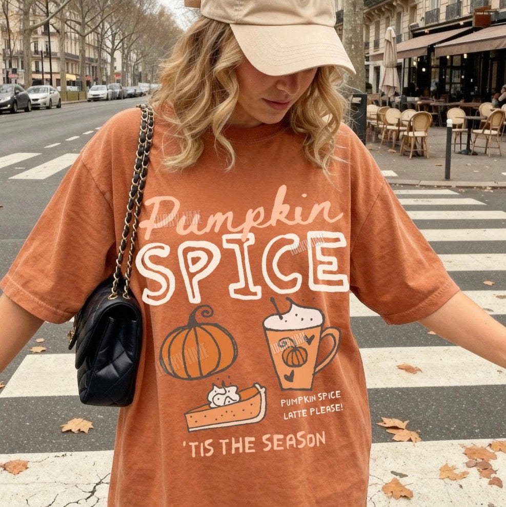 A woman wearing an oversized burnt orange graphic t-shirt with 'Pumpkin Spice' text and illustrations, paired with a beige baseball cap and a black quilted shoulder bag, walking on a city crosswalk.