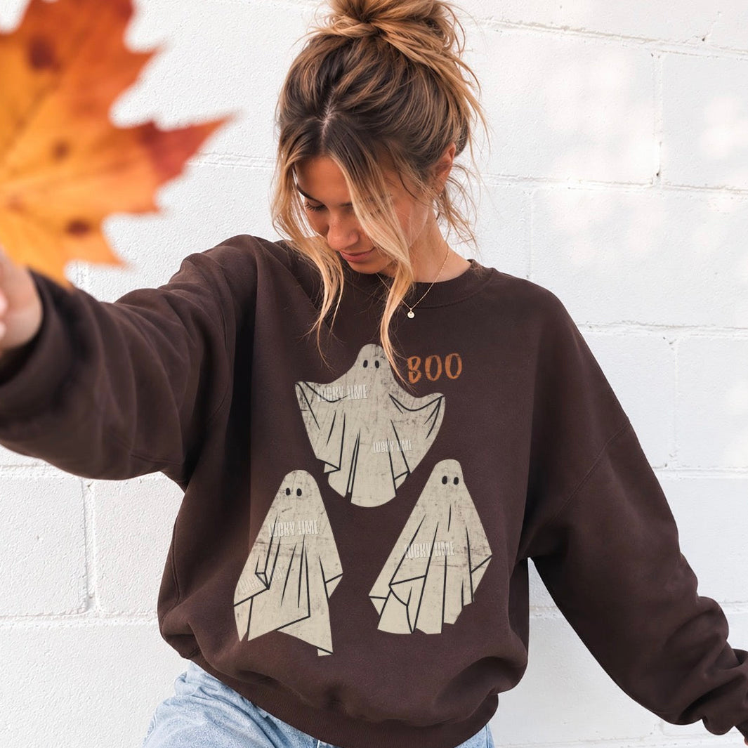 A woman wearing a dark brown crewneck sweatshirt with a graphic of three cream-colored ghosts and the word 'BOO' in orange. She is standing against a white brick wall, holding an autumn leaf in the foreground.