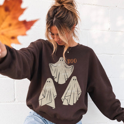 A woman wearing a dark brown crewneck sweatshirt with a graphic of three cream-colored ghosts and the word 'BOO' in orange. She is standing against a white brick wall, holding an autumn leaf in the foreground.