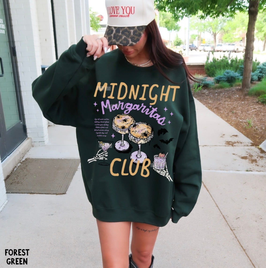 A woman wearing an oversized forest green sweatshirt with a colorful graphic that reads 'MIDNIGHT Margarita CLUB' and illustrations of donuts, a margarita glass, and a taco. She is standing outdoors on a sidewalk next to a building.
