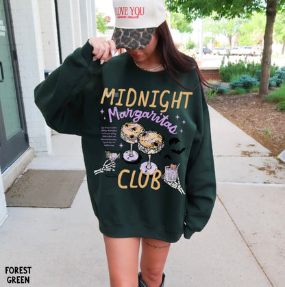 A woman wearing an oversized forest green sweatshirt with a colorful graphic that reads 'MIDNIGHT Margarita CLUB' and illustrations of donuts, a margarita glass, and a taco. She is standing outdoors on a sidewalk next to a building.