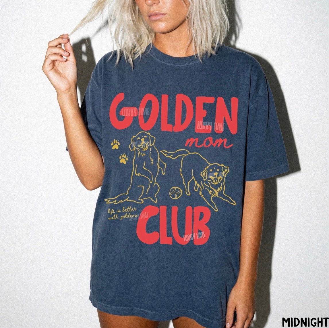 A woman wearing an oversized navy blue graphic t-shirt with 'GOLDEN mom CLUB' printed in red and yellow, featuring illustrations of two golden retrievers. The photo is taken against a plain white wall with a soft shadow.