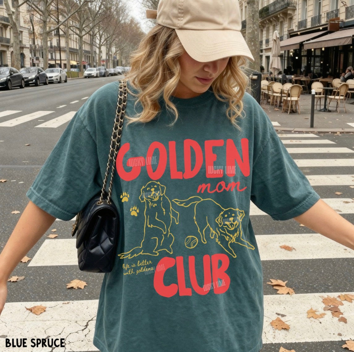 A woman wearing an oversized teal graphic T-shirt with red and yellow text reading 'Golden Mom Club' and dog illustrations, standing on a city crosswalk. She is also wearing a beige cap and carrying a black quilted shoulder bag.