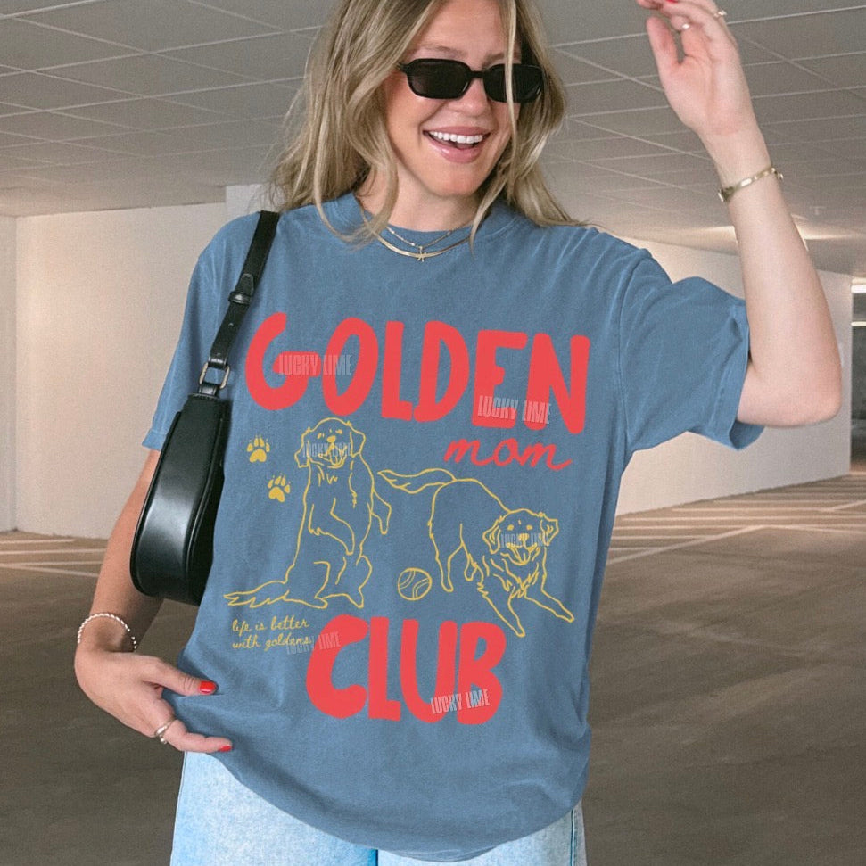 A woman wearing a blue graphic t-shirt with a red and yellow 'Golden Mom Club' print, styled with light blue jeans and a black shoulder bag in an indoor parking garage.
