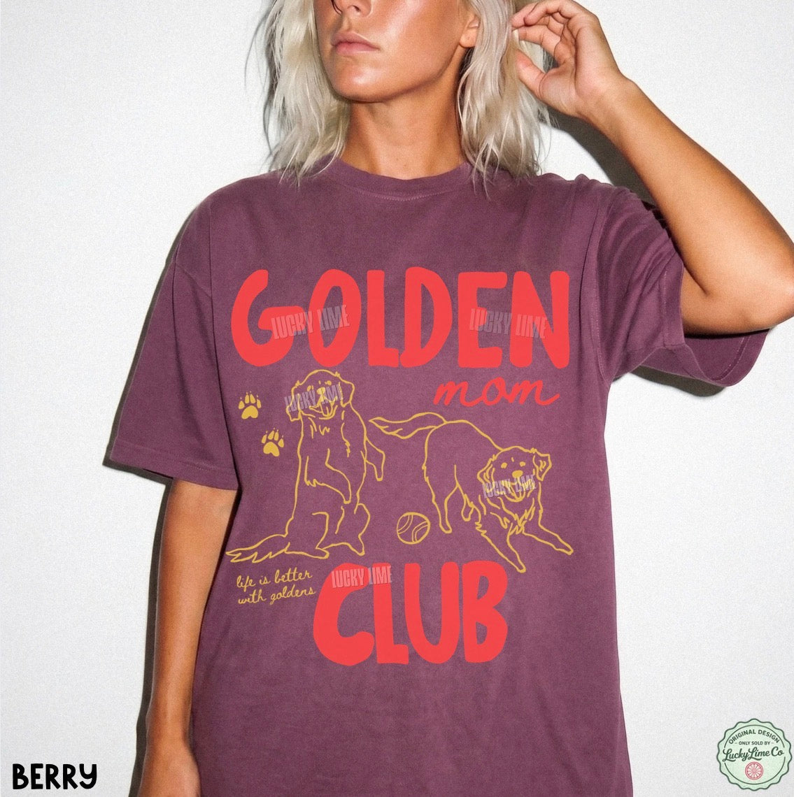 A woman wearing a berry purple graphic t-shirt with a yellow and red 'Golden Mom Club' dog design, shown against a plain white background.