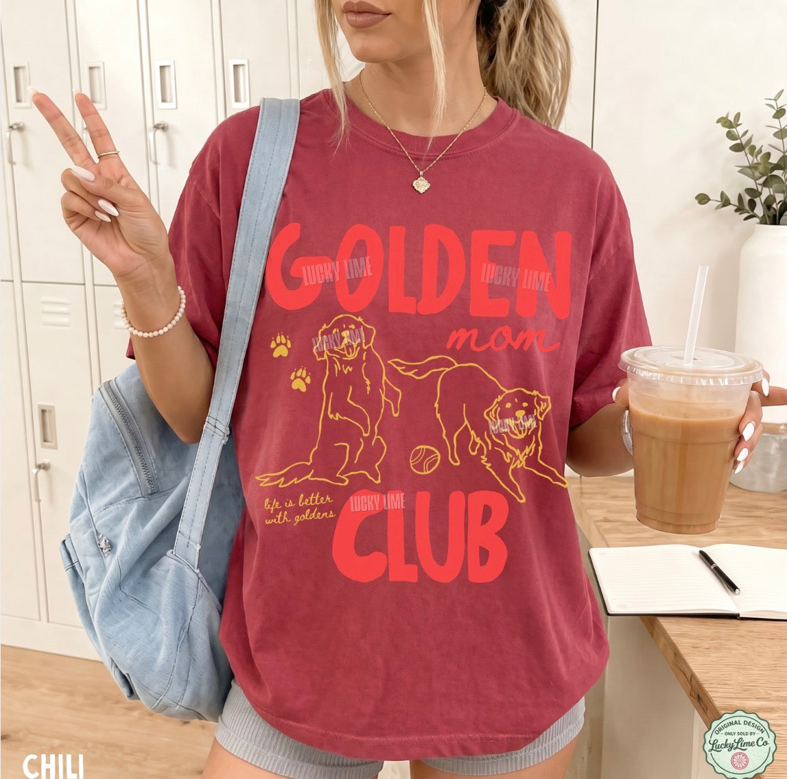A woman wearing a faded red graphic t-shirt with orange text reading 'GOLDEN MOM CLUB' and illustrations of two golden dogs. She is holding a light blue denim shoulder bag and a drink, standing in front of white lockers.
