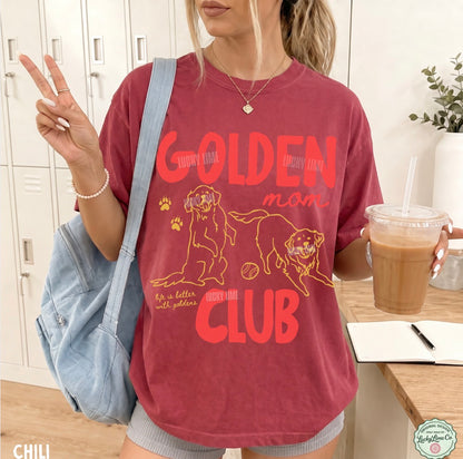 A woman wearing a faded red graphic t-shirt with orange text reading 'GOLDEN MOM CLUB' and illustrations of two golden dogs. She is holding a light blue denim shoulder bag and a drink, standing in front of white lockers.