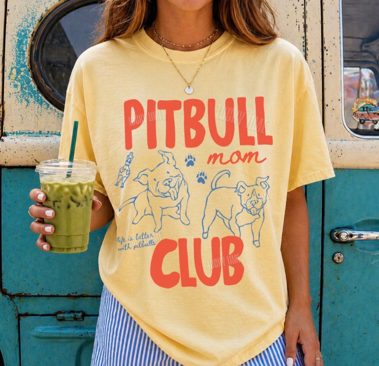 A woman wearing a pale yellow graphic t-shirt with 'PITBULL mom CLUB' printed in red and blue, holding an iced coffee in front of a rustic teal and cream wall.