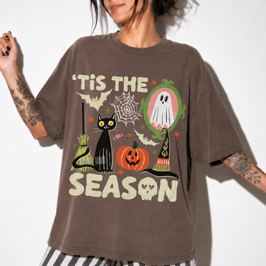 A person wearing an oversized espresso brown graphic T-shirt with Halloween-themed illustrations and text reading "'TIS THE SEASON," paired with black-and-white striped pants against a plain white background.