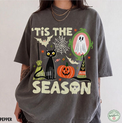A person wearing an oversized charcoal gray graphic T-shirt with Halloween-themed artwork including a black cat, pumpkin, ghost, broom, witch hat, bats, spiderwebs, and the text “'Tis the Season.”.