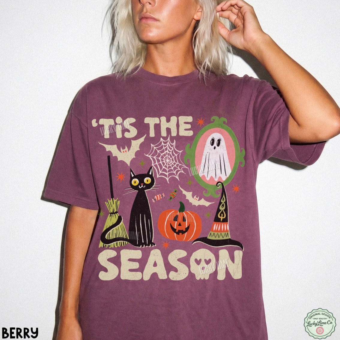 A woman wearing a berry purple graphic t-shirt with a Halloween design that reads "'TIS THE SEASON" featuring a black cat, broom, witch hat, pumpkin, ghost, and spiderweb. The photo is taken against a plain white wall.