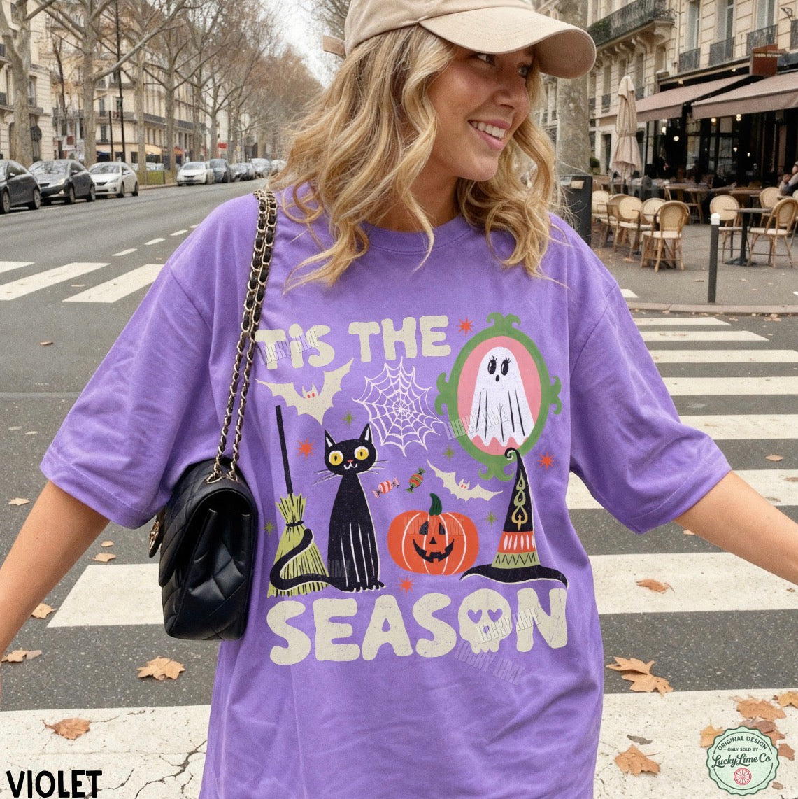 A woman wearing an oversized violet purple graphic t-shirt with a Halloween print, standing on a city street crosswalk. The t-shirt features a black cat, a jack-o'-lantern, a ghost, and a witch hat.