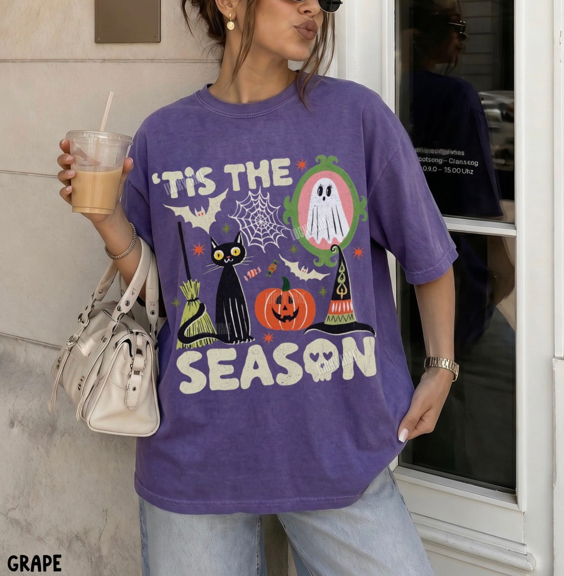 A woman wearing an oversized purple graphic T-shirt with Halloween-themed illustrations including a black cat, pumpkin, ghost, witch hat, spiderwebs, and the text “IT'S THE SEASON.” She is holding a drink and a white handbag, standing outdoors near a building entrance.