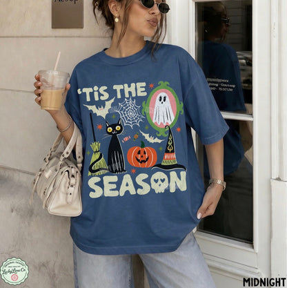 A woman wearing an oversized blue graphic T-shirt with a Halloween print featuring a black cat, pumpkin, ghost, witch hat, spiderwebs, and the text "TIS THE SEASON," photographed in a real doorway setting while holding an iced coffee.