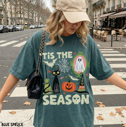 A woman wearing an oversized teal graphic t-shirt with a Halloween print, a beige baseball cap, and a black shoulder bag, standing on a city crosswalk.
