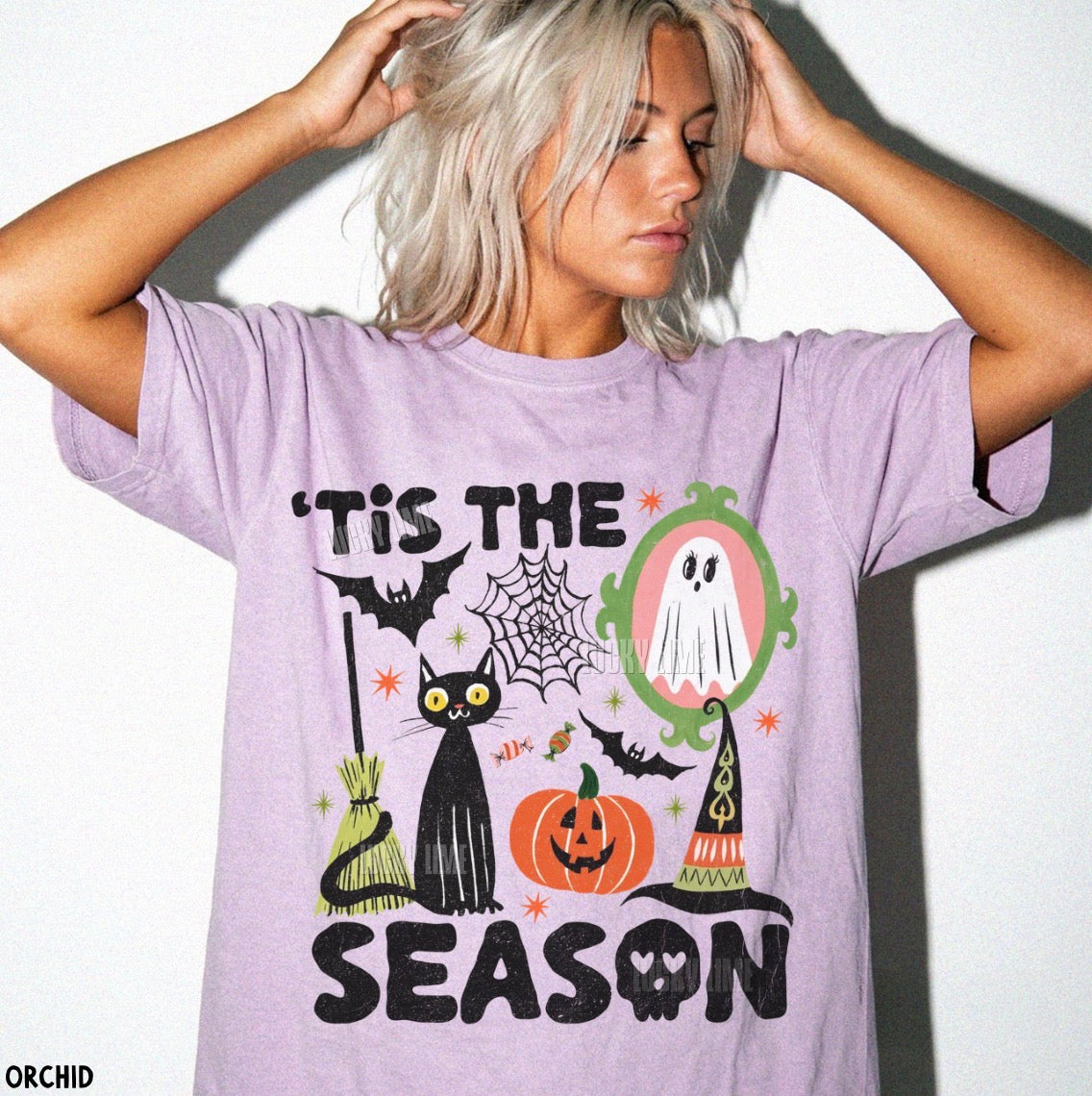 A woman with platinum blonde hair wearing a lavender graphic t-shirt with a Halloween print that reads "'TIS THE SEASON". The graphic features a black cat, pumpkin, witch hat, spiderwebs, and a ghost.