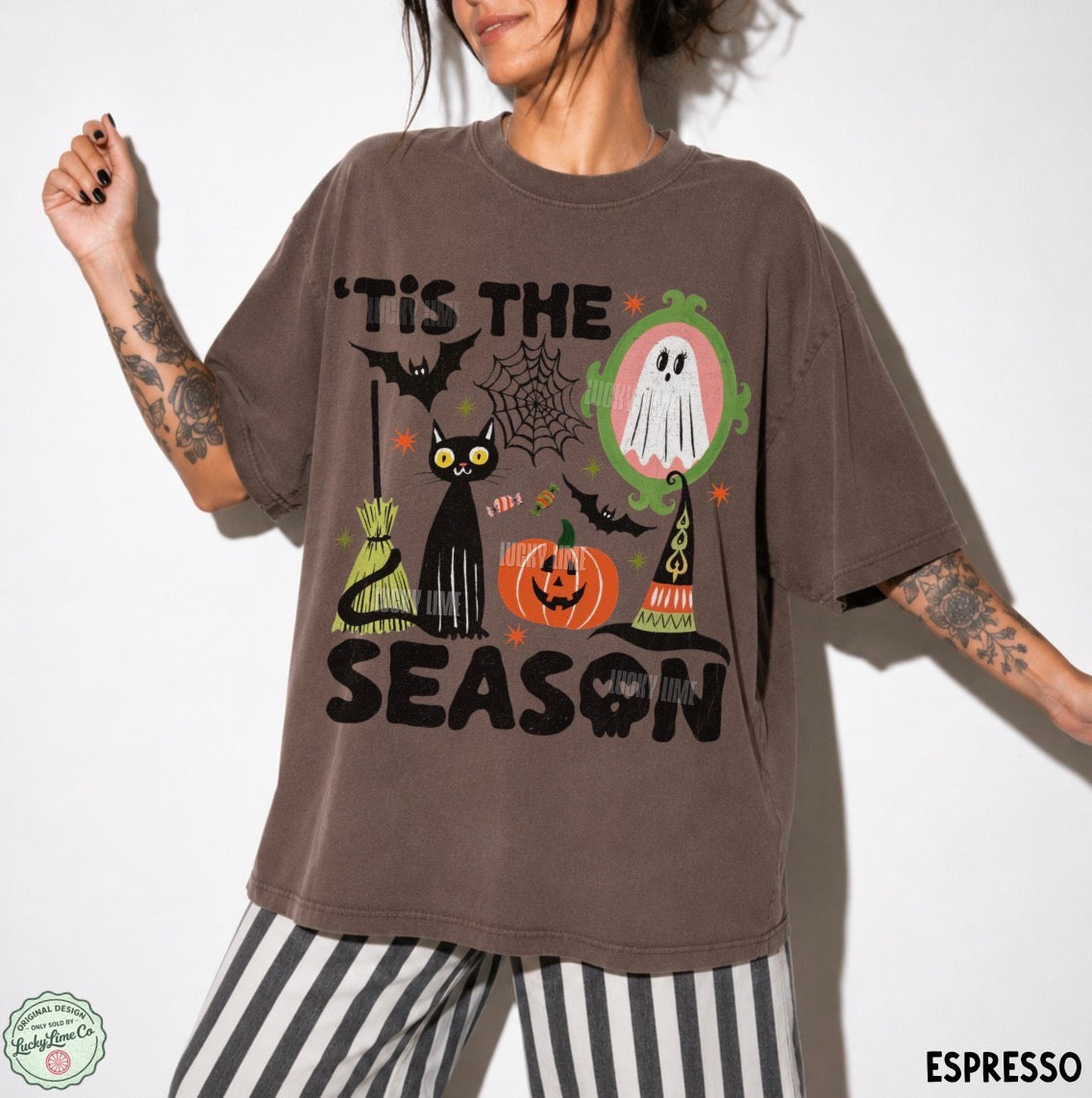 A woman wearing an oversized brown graphic t-shirt with a Halloween theme, paired with black and white striped pants. The t-shirt features a graphic print of a black cat, a jack-o'-lantern, a ghost in an oval mirror, and the text 'TIS THE SEASON'.