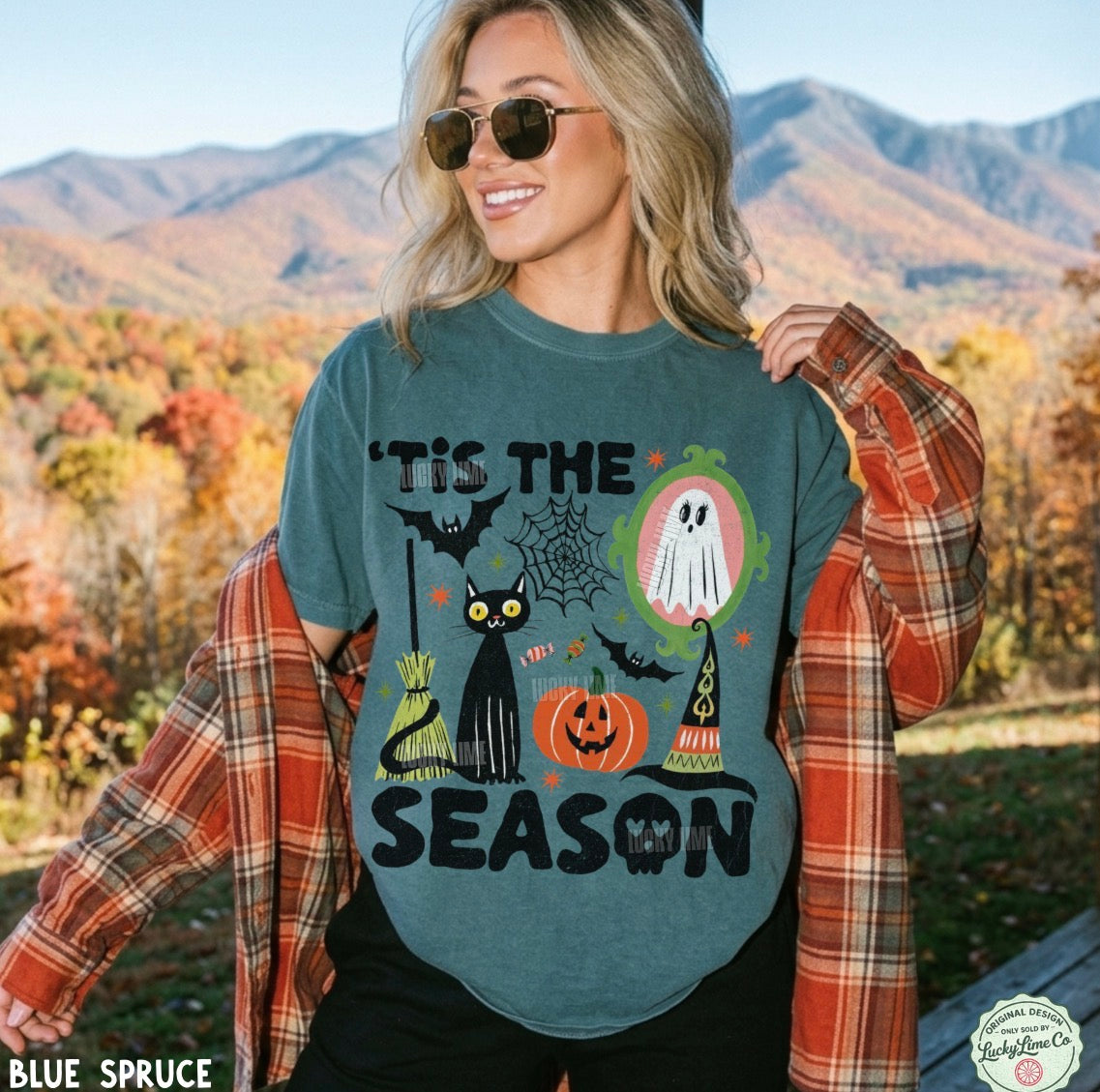 A woman outdoors wearing a slate blue graphic t-shirt with a Halloween design, layered with an orange plaid flannel shirt. The background shows autumn foliage and mountains.