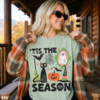 A woman wearing a sage green crewneck sweatshirt with a colorful Halloween graphic that reads 'TIS THE SEASON', styled with an orange and green plaid flannel shirt on a rustic wooden porch.