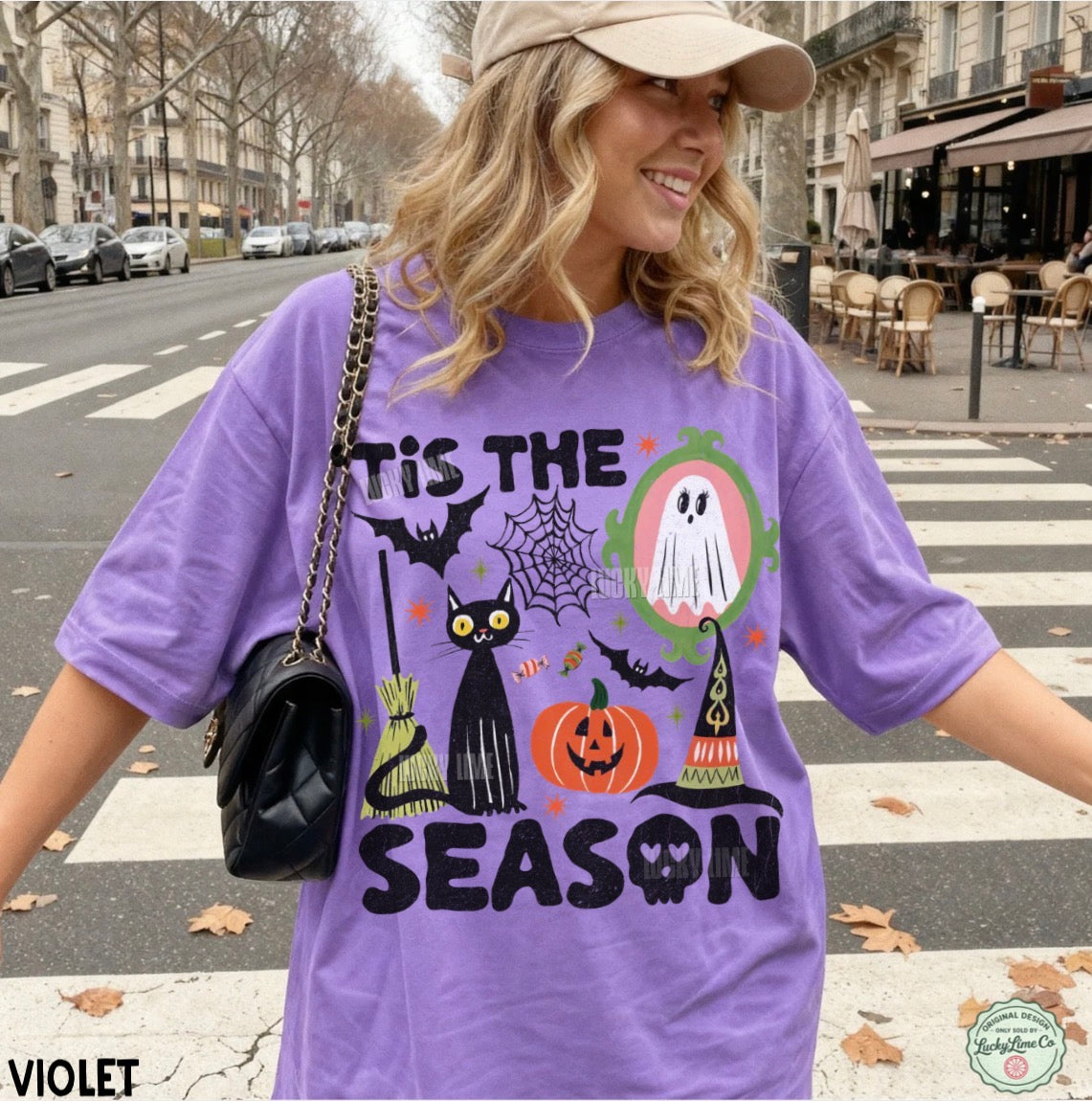 A woman wearing a lavender purple graphic t-shirt with a Halloween design that reads 'TIS THE SEASON' and features a black cat, pumpkin, witch hat, spiderwebs, and ghosts. She is standing on a city street crosswalk.