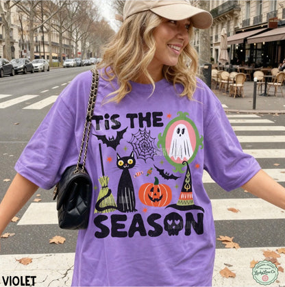A woman wearing a lavender purple graphic t-shirt with a Halloween design that reads 'TIS THE SEASON' and features a black cat, pumpkin, witch hat, spiderwebs, and ghosts. She is standing on a city street crosswalk.