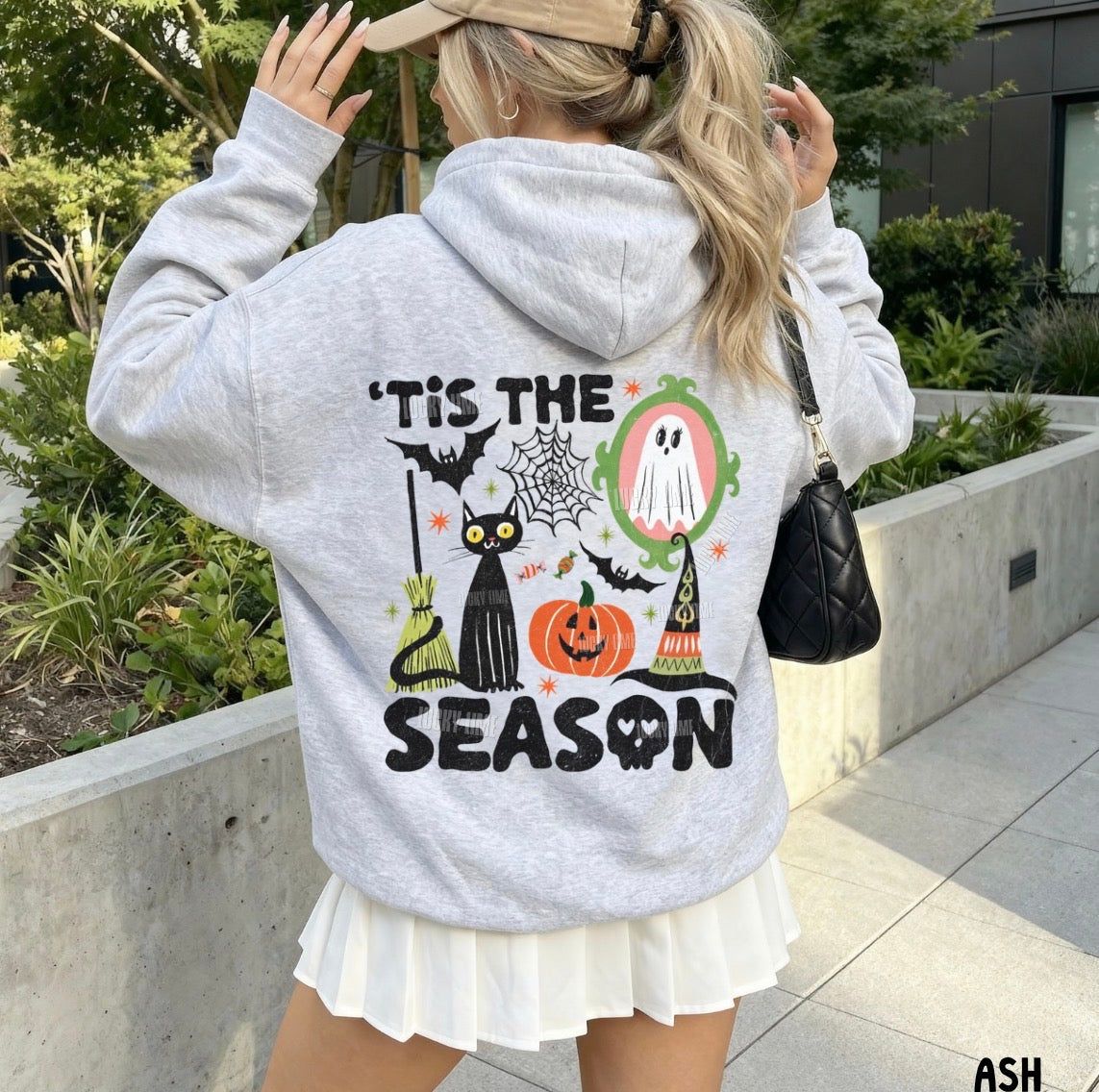A woman wearing a light gray heather hoodie with a large Halloween-themed graphic on the back, shown from behind in an outdoor setting. The graphic features the text "'TIS THE SEASON" with illustrations of a black cat, witch hat, spiderweb, ghost, and a jack-o'-lantern.