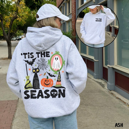A woman stands on a city sidewalk wearing a light gray hoodie from the back, which features a large Halloween graphic with a black cat, pumpkin, witch hat, spiderwebs, and the text “TIS THE SEASON.” An inset circle in the upper right shows the front of the hoodie with the text “TIS THE SEASON” printed on the chest.