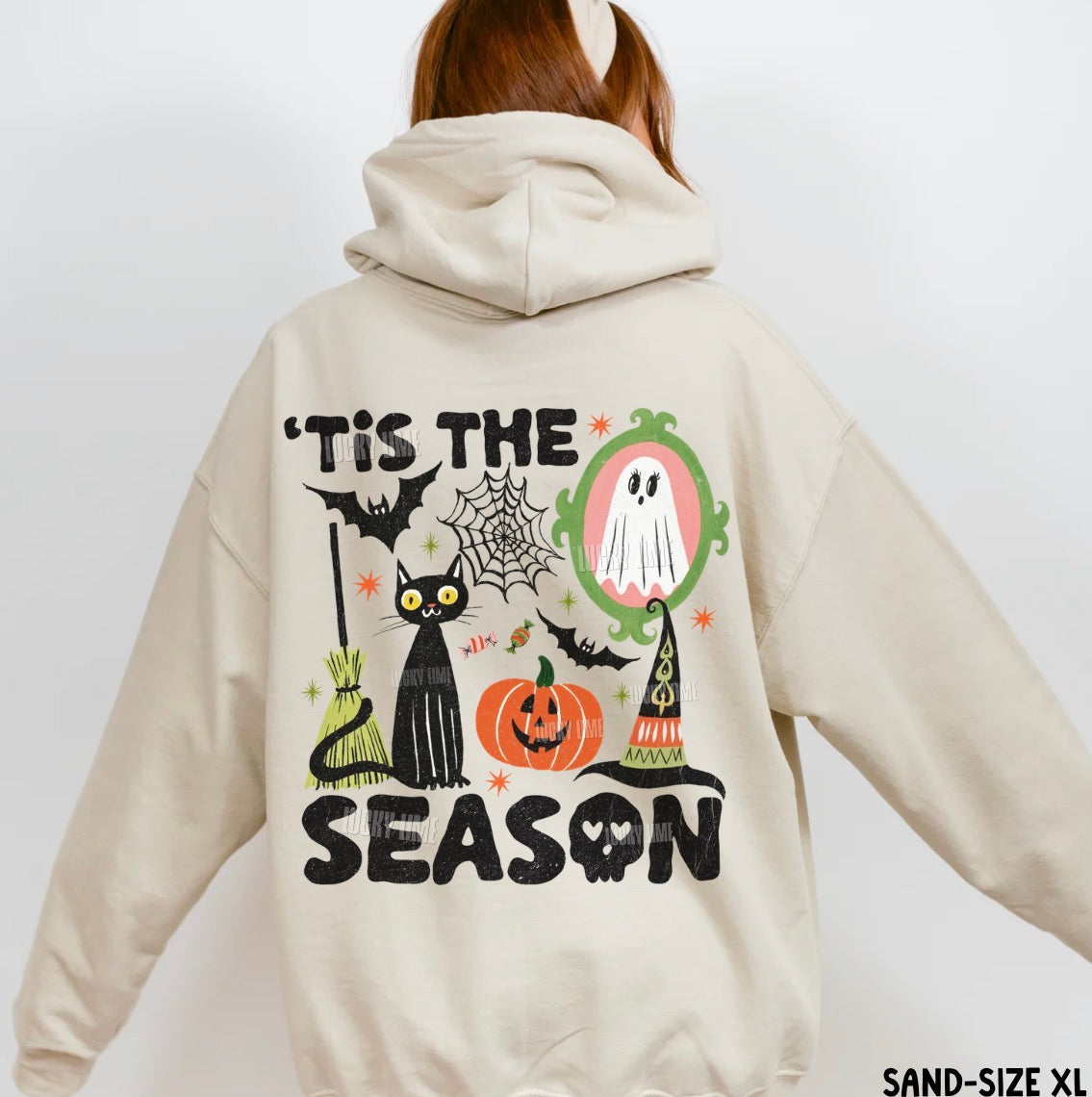A person shown from the back wearing a light beige hooded sweatshirt with a large Halloween graphic on the back. The graphic includes the text “'Tis the Season” with a black cat, pumpkin, ghost, witch hat, spiderweb, and bats.