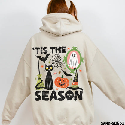 A person shown from the back wearing a light beige hooded sweatshirt with a large Halloween graphic on the back. The graphic includes the text “'Tis the Season” with a black cat, pumpkin, ghost, witch hat, spiderweb, and bats.