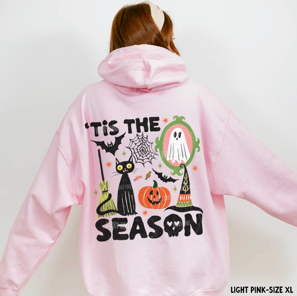 A person shown from the back wearing a light pink hoodie with a large Halloween graphic on the back. The graphic includes the text “tis THE SEASON,” a black cat, a ghost in a mirror, a witch hat, a pumpkin, bats, and spiderwebs.