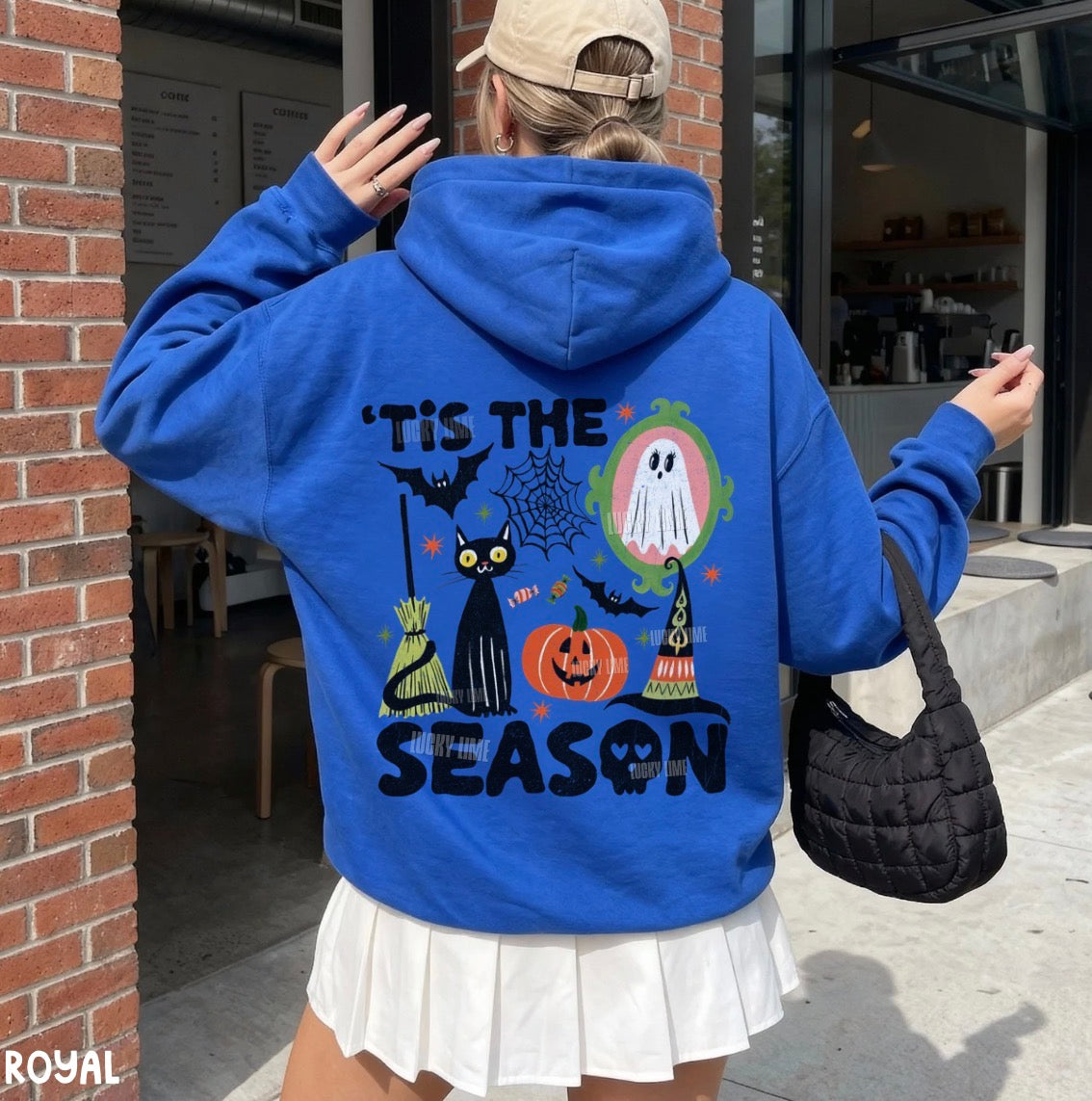 A woman wearing a bright blue hoodie with a large Halloween graphic on the back, shown from behind in an outdoor setting next to a brick wall. The graphic features a black cat, witch, pumpkin, ghost, and the text 'TIS THE SEASON'.