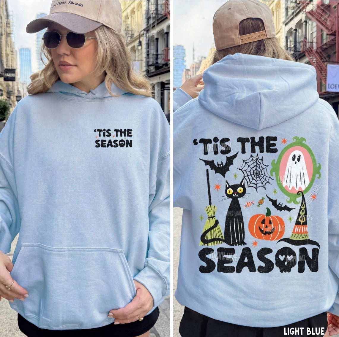 A woman modeling a light blue hoodie in an outdoor urban setting, shown from both the front and back. The front features the text 'TIS THE SEASON' in black on the left chest.