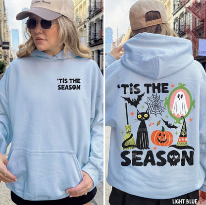 A woman modeling a light blue hoodie in an outdoor urban setting, shown from both the front and back. The front features the text 'TIS THE SEASON' in black on the left chest.