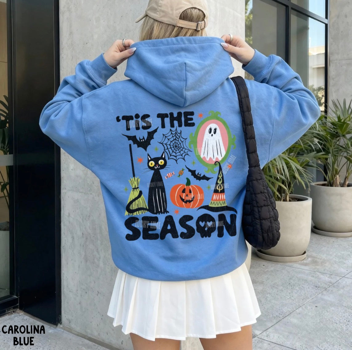 A person wearing a light blue hoodie with a large Halloween graphic on the back, including bats, a cat, spiderwebs, a ghost, pumpkins, and the text “'Tis the Season,” photographed outdoors near potted plants.