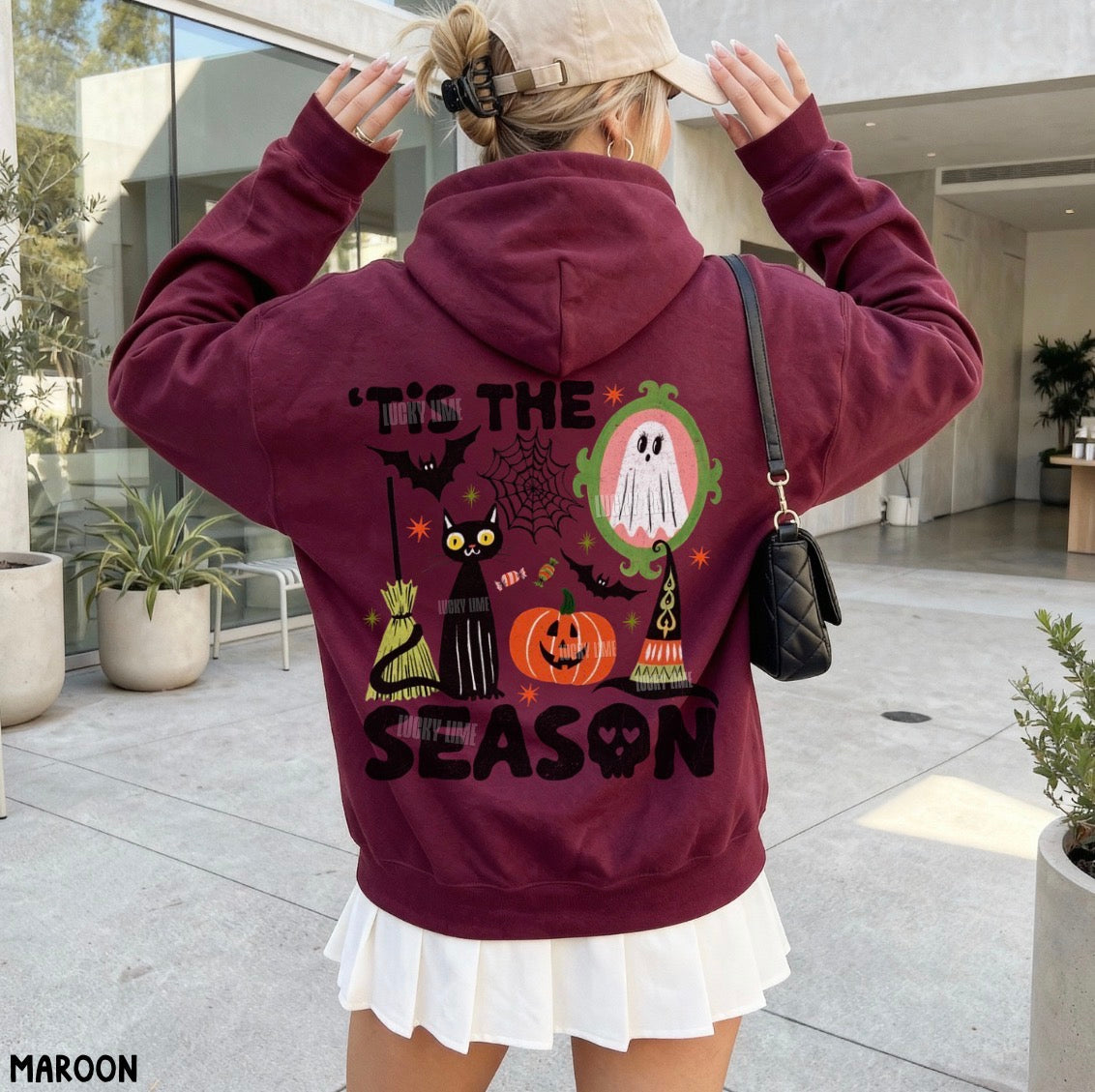 A woman wearing a maroon hoodie with a large Halloween graphic on the back, including a ghost, pumpkin, cat, witch, and the text "Tie the Season". She is wearing a beige cap and a white pleated skirt in an outdoor patio setting.