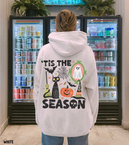 A person viewed from behind wearing a white hoodie with a large Halloween graphic and the text “'Tis the Season,” standing in front of two beverage refrigerators.