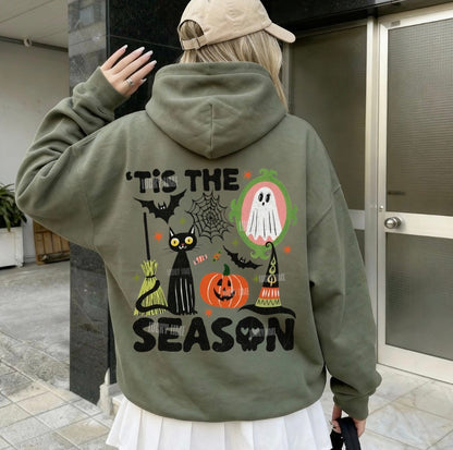 A person shown from behind wearing an olive green hoodie with a colorful Halloween graphic on the back that reads 'TIS THE SEASON'. The hoodie features illustrations of a black cat, jack-o'-lantern, ghost, witch hat, spiderwebs, and bats.
