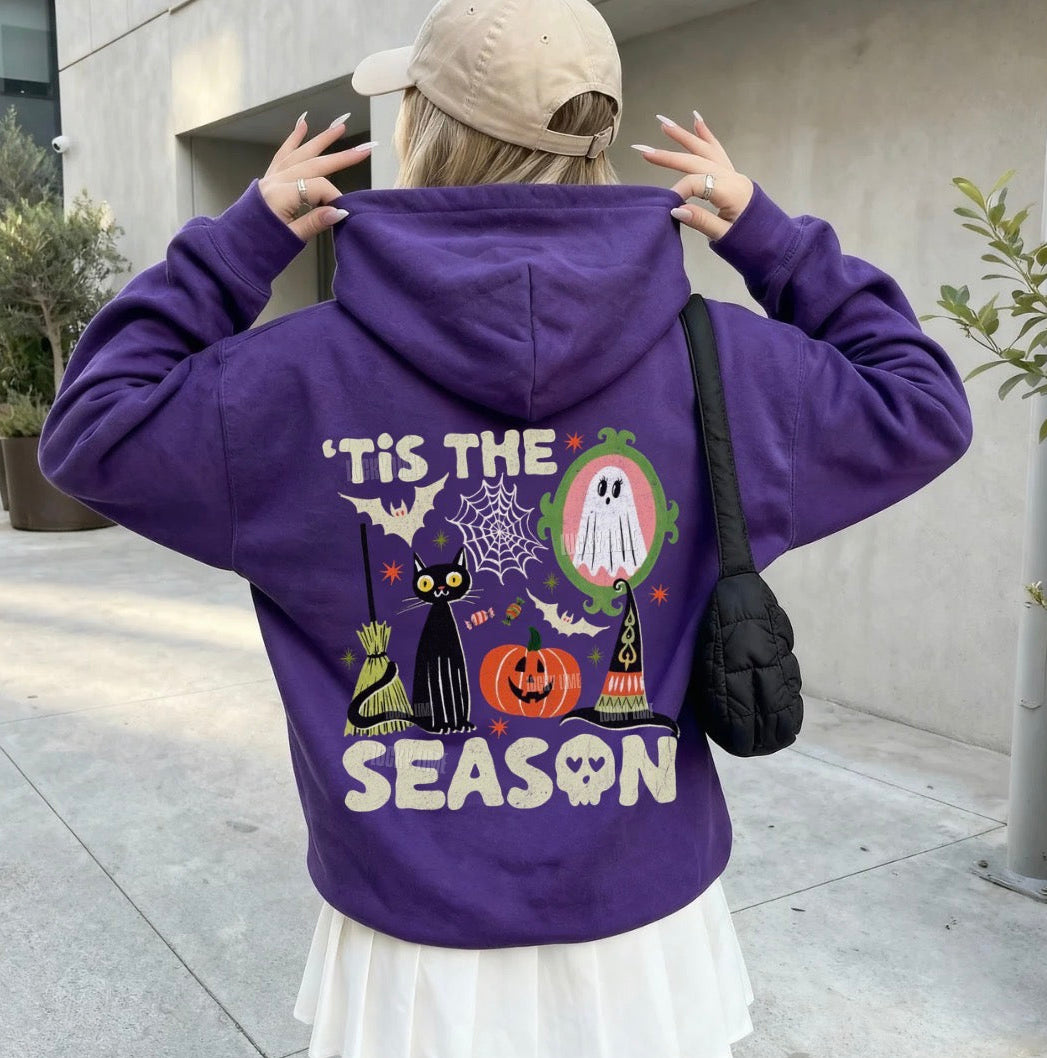 A woman wearing a purple hoodie with a Halloween-themed graphic on the back, featuring a black cat, witch, ghost, pumpkin, and the text 'Tis the Season'. She is wearing a beige cap and a white pleated skirt, standing outdoors on a concrete walkway.