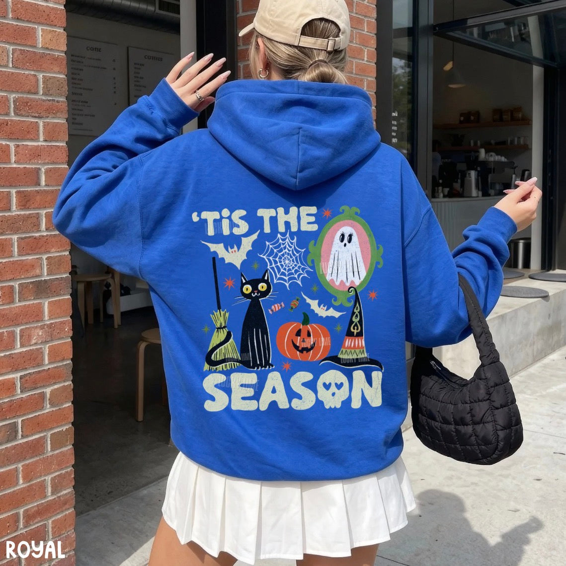 A woman viewed from behind wearing a bright blue hoodie with a large Halloween-themed graphic on the back, including a black cat, pumpkin, ghost, broom, witch hat, spiderweb, and the text “'Tis the Season.” She is also wearing a beige cap and a white pleated skirt, standing outdoors near a brick building.