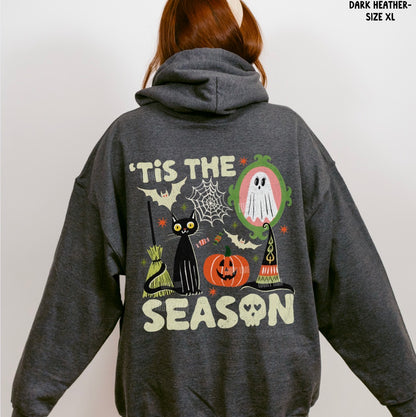 A person shown from the back wearing a dark heather gray hoodie with a large Halloween-themed graphic on the back. The design includes the text “'Tis the Season,” a black cat, a jack-o'-lantern, a ghost, bats, a broom, and a witch hat.