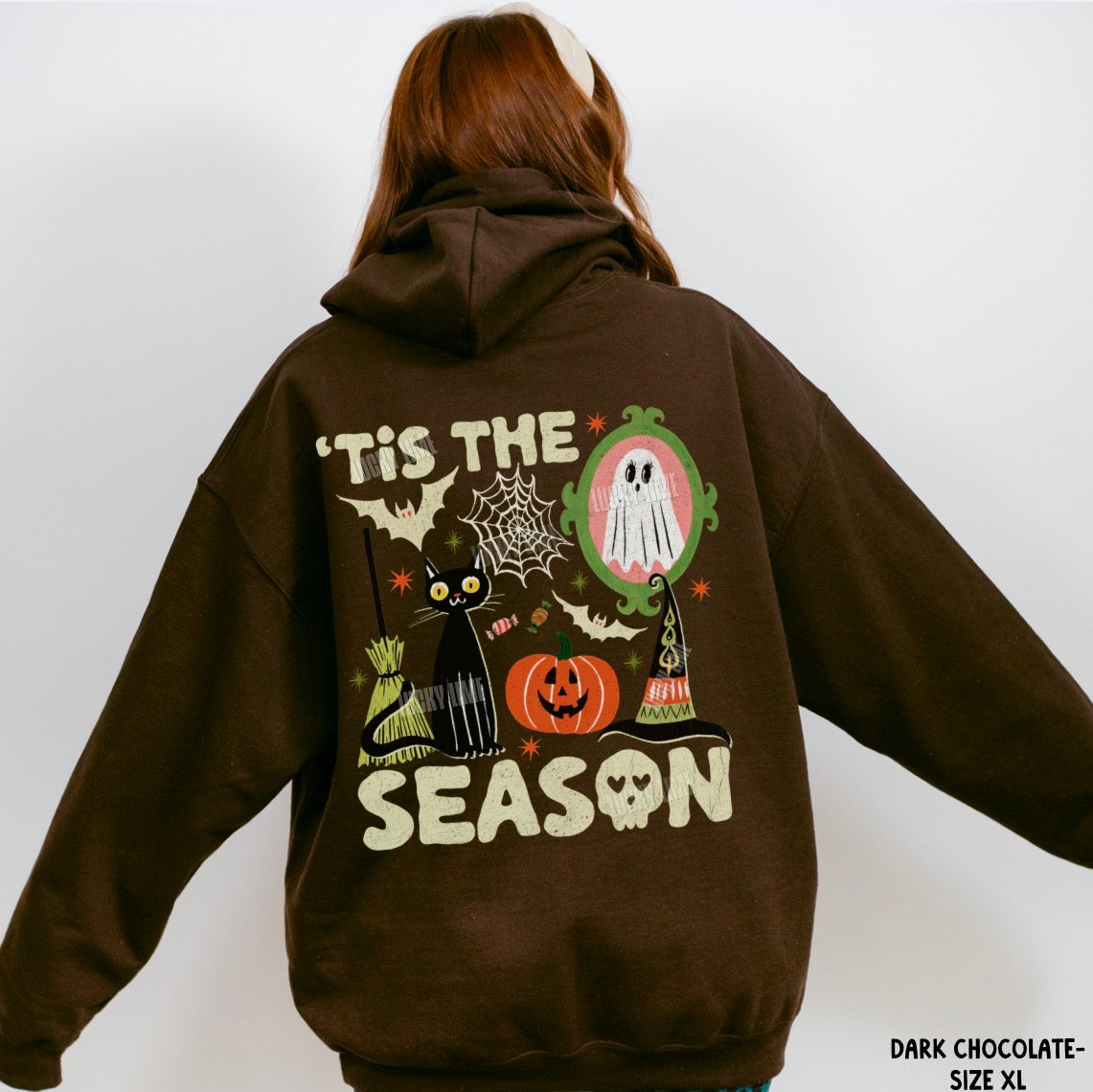 A person shown from the back wearing a dark chocolate brown hoodie with a large Halloween graphic on the back. The graphic features the text 'IT'S THE SEASON' in cream, a black cat, a jack-o'-lantern, a ghost in an oval mirror frame, a witch's hat, bats, and cobwebs.