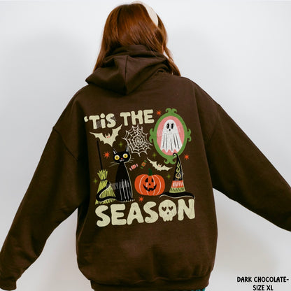 A person shown from the back wearing a dark chocolate brown hoodie with a large Halloween graphic on the back. The graphic features the text 'IT'S THE SEASON' in cream, a black cat, a jack-o'-lantern, a ghost in an oval mirror frame, a witch's hat, bats, and cobwebs.