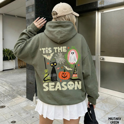 A woman from behind wearing an olive green hoodie with a large Halloween graphic on the back that reads "'TIS THE SEASON" with a ghost, cat, pumpkin, witch hat, bats, and spiderweb. She is wearing a beige baseball cap and a white pleated skirt, standing outside near a building entrance.